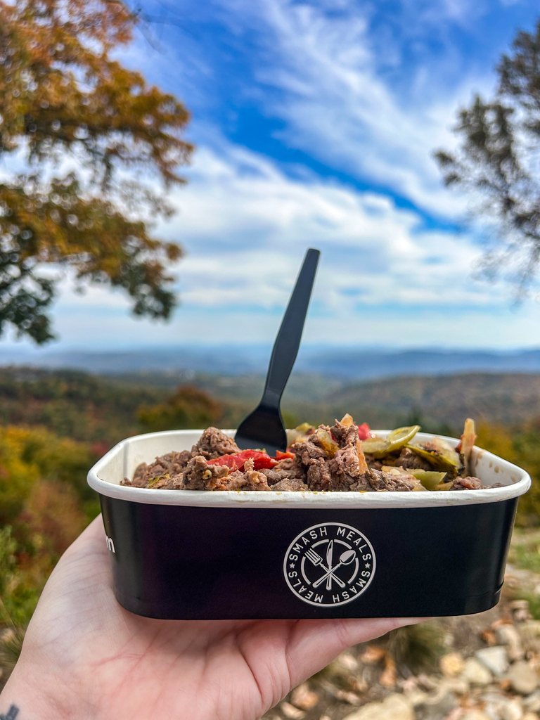 A hand holds a SmashMeals meal with a black fork in front of tree-covered hills.